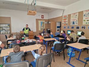 na zdjęciu policjant stojący przed dziećmi siedzącymi w ławkach szkolnych