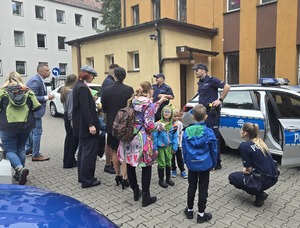 na zdjęciu radiowóz policyjny, przy nim dwóch policjantów, policjantka i zgromadzone osoby