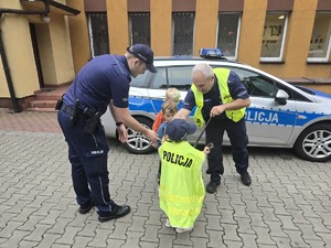 na zdjęciu radiowóz policyjny, przy nim dwóch policjantów i trójka dzieci, jedno z nich w czapce i kamizelce policyjnej