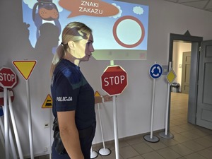 na zdjęciu policjantka, obok znaki drogowe, w dłoni trzyma znak stop