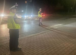 na zdjęciu dwóch policjantów drogówki, jeden z latarką w dłoni, obok radiowóz