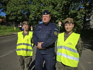 na zdjęciu dwie wojskowe w żółtych kamizelkach, pomiędzy nimi policjant
