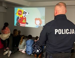 na zdjęciu dzieci oglądają film o bezpieczeństwie, obok stoi policjant, w budynku komisariatu