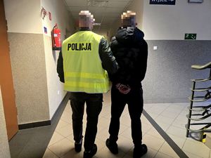 nieumundurowany policjant w kamizelce z napisem Policja trzyma za ramię zatrzymanego mężczyzne w kajdankach