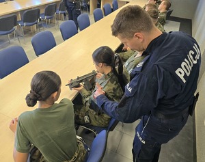 na zdjęciu uczniowie w strojach moro siedzą przy stołach, oglądają broń służbową, przy nich policjant