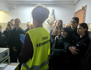 na zdjęciu techniczka kryminalistyki w żółtej kamizelce, przy niej licealiści w czarnych strojach