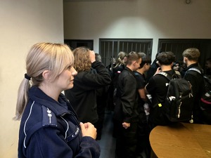 na zdjęciu policjantka, obok licealiści przy kratach, w pomieszczeniu dla zatrzymanych osób