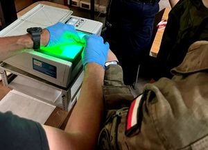 na zdjęciu dłonie w niebieskich rękawiczkach i dłoń ucznia w stroju moro na urządzeniu do pobierania odcisków palców