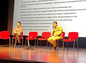 na zdjęciu na scenie dwie osoby prowadzące debatę, za nimi wyświetlony slajd dotyczący uzależnień
