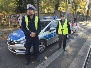 na zdjęciu policjant i strażniczka miejska przy radiowozie obok cmentarza