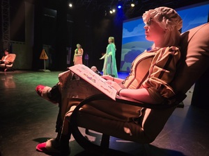 na zdjęciu aktorzy na scenie w trakcie przedstawienia, dziewczyna siedząca na fotelu trzyma w dłoniach książkę i mikrofon, za nią dwie dziewczyny