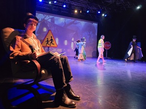 chłopiec w kapeluszu siedzi na fotelu, na kolanach ma książkę a w dłoni mikrofon, obok aktorzy przebrani za postacie z bajki