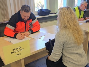 przy stole kobieta i ratownik medyczny, na stole kartka na niej napis zespół medyczny