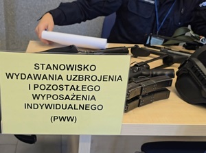 na zdjęciu na stole kartka na niej napis stanowisko wydawania uzbrojenia i pozostałego wyposażenia indywidualnego, za nią leżąca na stole broń obok policjant