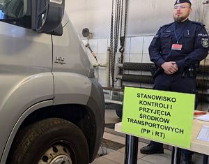 na zdjęciu na stole żółta kartka na niej napis stanowisko kontroli przyjęcia środków transportu, za stolikiem policjant, obok samochód szarego koloru