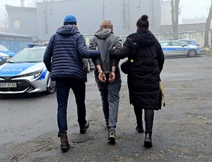 zatrzymany mężczyzna prowadzony przez policjanta i policjantkę na parkingu komendy