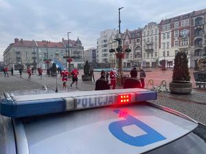 na zdjęciu radiowóz na rynku, obok ludzie biegnący w mikołajowych strojach