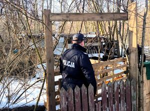 na zdjęciu policjant znajdujący się obok ogrodzenia i drzew za którymi stoi niewielki budynek