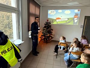 na zdjęciu policjant obok siedzące na krzesłach dzieci w sali, przed nimi wyświetlane slajdu dotyczące bezpieczeństwa