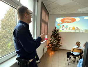 na zdjęciu policjant obok siedzące na krzesłach dzieci w sali, przed nimi wyświetlane slajdu dotyczące bezpieczeństwa