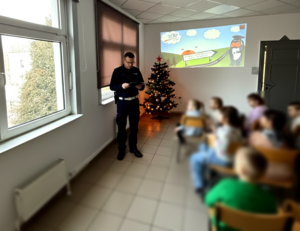na zdjęciu policjant obok siedzące na krzesłach dzieci w sali, przed nimi wyświetlane slajdu dotyczące bezpieczeństwa
