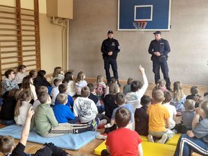dwóch policjantów stojących przed dziećmi siedzącymi w sali gimnastycznej