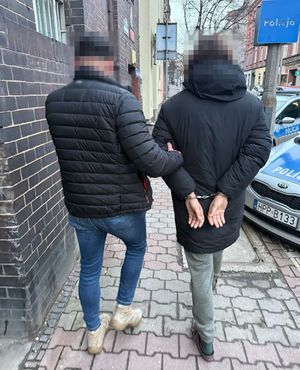 na zdjęciu zatrzymany mężczyzna w kajdankach zapiętych z tyłu prowadzony przez nieumundurowanego policjanta