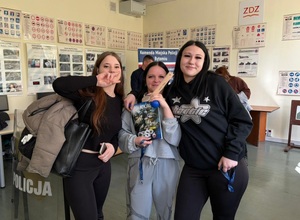 na zdjęciu trzy dziewczyny stojące obok siebie i pozujące do zdjęcia, dziewczyna w środku trzyma w dłoniach gazetę policyjną