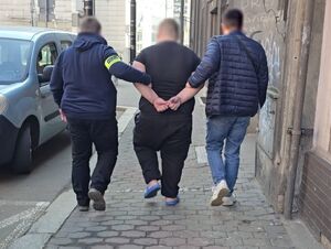 dwóch nieumundurowanych policjantów prowadzi zatrzymanego mężczyznę w kajdankach przed budynkiem prokuratury