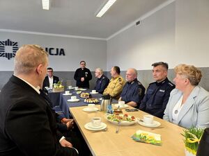 kapelan stojący przy stole i siedzący przy stołach policjanci i pracownicy cywilni