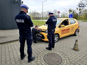 widzimy dwóch policjantów drogówki, przed nimi chłopak przy samochodzie do jazdy egzaminacyjnej podnoszący pokrywę silnika pojazdu