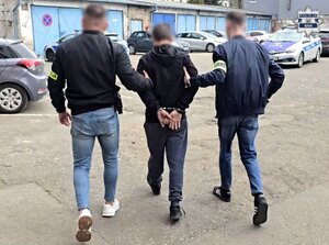 na parkingu policyjnym dwóch nieumundurowanych policjantów prowadzi zatrzymanego mężczyznę, zatrzymany ma dłonie w kajdankach zapiętych z tyłu