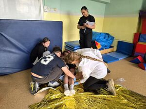 dwie dziewczyny i chłopak bandażują nogi leżącemu na podłodze w sali chłopakowi obok stoi kobieta