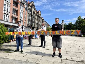 dwóch młodych mężczyzn trzyma napisy żółto pomarańczowe liter układają się w wyrazy Happening Czytelniczy