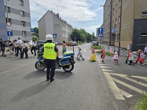 policjant przy motocyklu na środku jezdni, obok przechodzą dzieci w kolorowych strojach