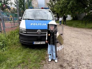 dziewczynka stoją ca przed radiowozem, ubrana w kask trzyma w ręku pałkę i tarczę policyjną