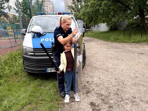policjantka i dziewczynka stojąca przed radiowozem, trzyma w ręku pałkę. policjantka podaje jej tarczę policyjną