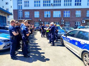 policjanci ustawieni w rzędach, trzymają w dłoniach notatniki, są na przeciwko kilku innych policjantów, obok radiowozy