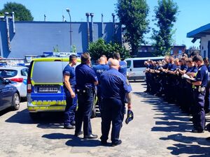 policjanci ustawieni w rzędach, trzymają w dłoniach notatniki, są na przeciwko kilku innych policjantów, obok radiowozy