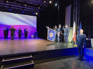 dowódca uroczystości, obok na scenie poczet sztandarowy, policjanci salutują, obok występ policyjnej orkiestry, zanimi wyświetlona flaga Polski