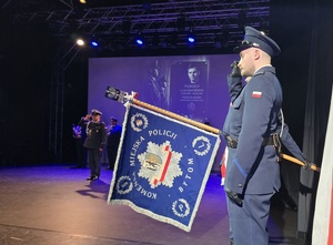 poczet sztandarowy, orkiestra na scenie, policjanci salutują, za nimi wyświetlona grafika, na niej policjanci i pomnik Marka Sienickiego