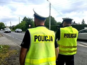 policjant w trakcie pomiaru prędkości, trzyma urządzenie w dłoniach przy twarzy, obok niego kolejny policjant