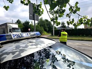 policjant w trakcie pomiaru prędkości, trzyma urządzenie w dłoniach przy twarzy obok niego radiowóz