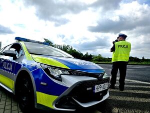 policjant w trakcie pomiaru prędkości, trzyma urządzenie w dłoniach przy twarzy, obok niego radiowóz