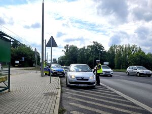 policjant w trakcie kontroli pojazdu, stoi przy oknie kierowcy pojazdu srebrnego koloru, za nim radiowóz i kolejny policjant