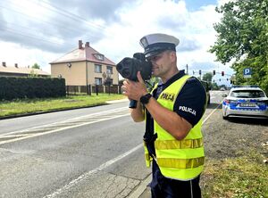 policjant w trakcie pomiaru prędkości, trzyma urządzenie w dłoniach przy twarzy, obok niego radiowóz
