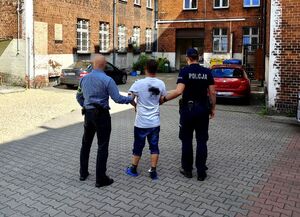 dwóch policjantów, jeden nieumundurowany trzyma zatrzymanego mężczyznę pod ramię, mężczyzna ma na sobie kajdanki zespolone