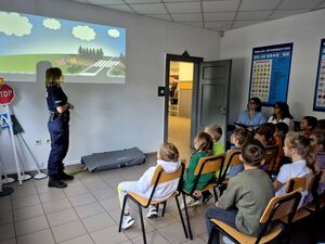 dzieci siedzące w salina krzesłach, przed nimi policjantka, za nią slajd przedstawiający drogę