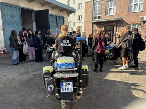 policjant, policjantka, dwa motocykle policyjne i licealiści zgromadzeni na parkingu, na jednym z motocykli siedzi uczennica klasy mundurowej