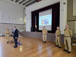 na sali mężczyzna w garniturze stojący przy mikrofonie obok strażak, za nimi trzech strażaków